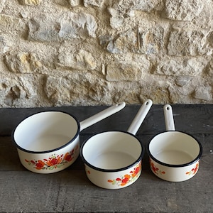 Op de afbeelding: Drie vintage emaille sauspannen met witte binnenkanten, zwarte randen en lange witte handvatten. De crèmekleurige buitenkanten hebben een bloemenontwerp in rood, oranje en groen. De pannen staan op een verweerd houten oppervlak.