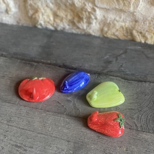 Op de afbeelding: Een verzameling keramische groente-vormige decoratieve items. De set bevat een rode tomaat, een blauwe paprika, een groene paprika en een rode aardbei. De items zijn gerangschikt op een verweerd houten oppervlak.