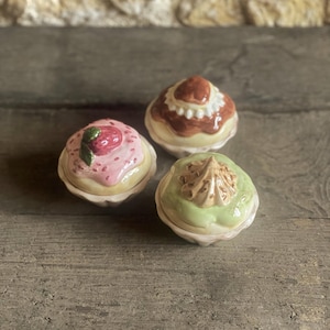 Op de afbeelding: Drie keramische cupcake-vormige sieradendoosjes. Eén is roze met een aardbei, een andere heeft bruine glazuur en de derde is groen met een wervelontwerp. De doosjes staan op een verweerd houten oppervlak.