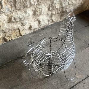 Op de afbeelding: Een zilverkleurige draadsculptuur van een vogel, mogelijk een kip, met een afgerond lichaam en een lange nek. De sculptuur is gemaakt van dunne metalen draden, waardoor een open, kooi-achtige structuur ontstaat. De vogel rust op een houten oppervlak.
