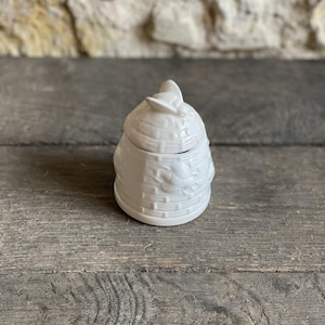 Può includere: Un piccolo contenitore in ceramica bianca con coperchio, a forma di alveare. Il contenitore ha una superficie strutturata e un piccolo elemento decorativo sulla parte superiore. Su una superficie di legno rustico, con un muro di pietra sullo sfondo.