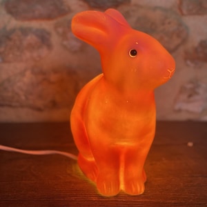 Puede incluir: Una lámpara brillante con forma de conejo naranja y acabado liso y translúcido. La lámpara presenta orejas erguidas, un pequeño ojo oscuro y un detalle de nariz moldeado. Un fino cable de alimentación blanco sale de la base de la figura iluminada.