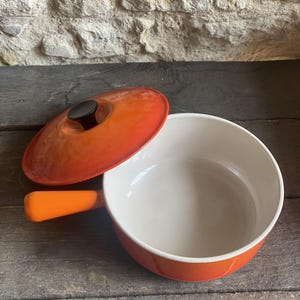 Op de afbeelding: Een ronde, oranje geëmailleerde gietijzeren steelpan met een bijpassend deksel voorzien van een zwarte knop. De binnenkant van de pan is crèmekleurig en heeft een enkele, lange, oranje steel die aan de zijkant van de pan uitsteekt.