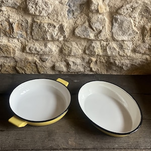 May include: Two oval white enamel baking dishes with black rims and yellow handles. They sit on a weathered wooden surface against a textured stone wall. One dish has handles. The dishes are for baking.