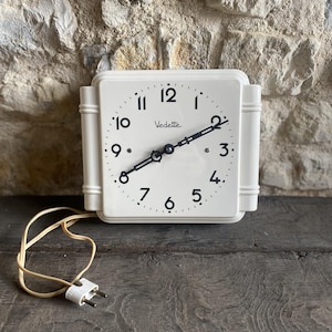 May include: A square, off-white Vedette clock with black numerals and hands. The clock has a cream-colored cord and plug. The clock is mounted on a wall with a stone texture.