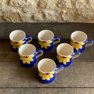 Op de afbeelding: Een set van zes keramische mokken met een felblauwe achtergrond en een handgeschilderd geel zonnebloemmotief aan de buitenkant. Elke mok heeft een witte binnenkant, een gebogen handvat en een afgeronde basis, gerangschikt in een groep.