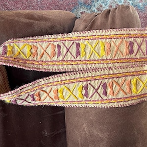 May include: A woven beige fabric strap with a geometric pattern of yellow, orange, and purple embroidery. The strap is folded in half and has a frayed edge.