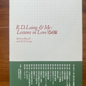 May include: A paperback book titled "R.D. Laing & Me: Lessons in Love" by Roberta Russell with R.D. Laing. The cover is off-white with text in black and red. Japanese text is at the top. The book's spine and back cover are green.
