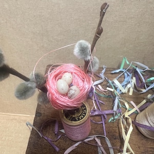 May include: A pink thread spool with a pink yarn nest on top, containing three speckled white Easter eggs. The spool is labeled "Cotton Twist" and is surrounded by colorful paper streamers.