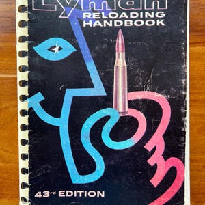 Puede incluir: Un manual de recarga Lyman vintage, 43ª edición, con encuadernación en espiral. La portada es negra con un diseño de rostro estilizado en azul y rosa, y un gráfico de bala. El título está en blanco y azul.