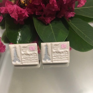May include: A pair of white ceramic earrings with a pink and gray Eiffel Tower design. The earrings are shaped like postcards.