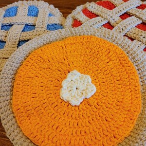 Peut inclure: Plusieurs articles faits à la main sur le thème de la tarte au crochet. L'image présente une tarte ronde orange avec un accent blanc en forme de fleur et deux dessous de plat en forme de tarte avec des croûtes en treillis bleues et rouges.
