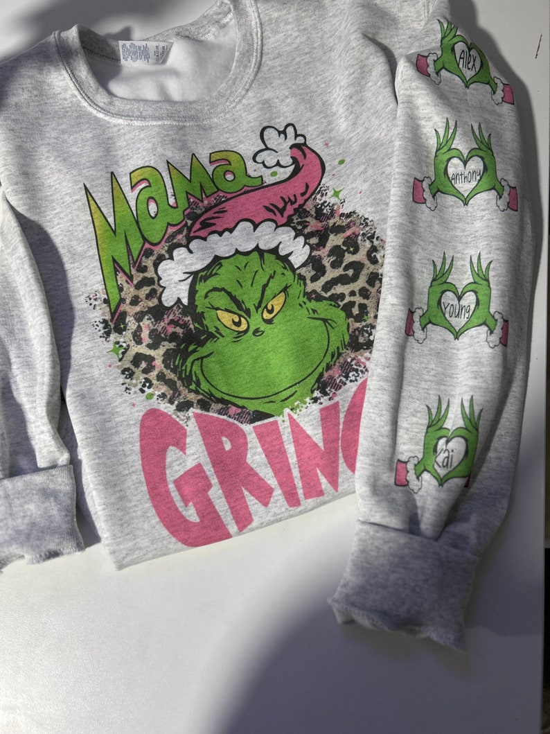 May include: Gray sweatshirt with a pink "Mama Grinch" graphic featuring the Grinch wearing a Santa hat and a leopard print background. The sleeves have pink hearts with the names "Alex", "Anthony", "Young", and "Kai" written inside.
