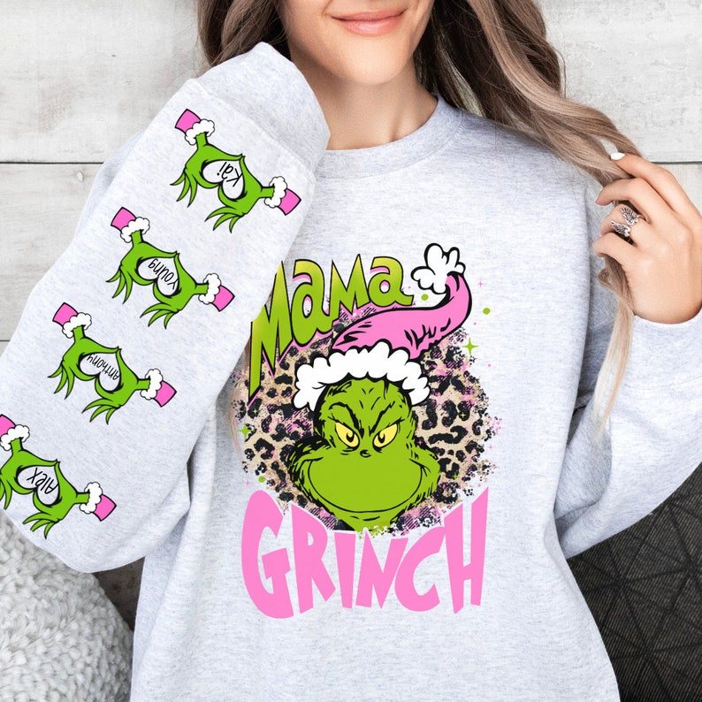 May include: A gray sweatshirt with a green Grinch character wearing a pink Santa hat on the front. The Grinch is surrounded by a leopard print design. The text "Mama Grinch" is written in pink letters above the Grinch. The sleeves of the sweatshirt have green Grinch hands with pink hearts and the words "Irey", "Bunox", "Aviomah", and "Xriv" written on them.