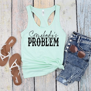 May include: A light green tank top with the text "Somebody's PROBLEM" in black lettering. The tank top is laying on a wooden surface with a pair of brown sandals and a pair of blue jeans.