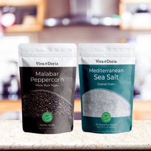 May include: Two pouches of spices and seasonings. The pouch on the left is black with white text and contains whole black peppercorns. The pouch on the right is teal with white text and contains coarse sea salt. Both pouches have a green logo with the text "Viva Doria" and a green "Natural" seal.