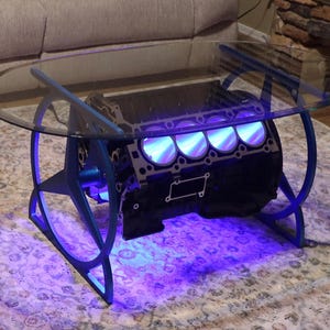 May include: A unique coffee table featuring a glass top supported by a blue metal frame. The base is a black engine block with illuminated cylinders, creating a striking industrial design. The table is approximately 36 inches in diameter.