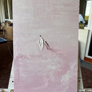 Pode incluir: Pintura abstrata em tela com fundo rosa texturizado. Uma forma branca em forma de pena e uma mancha de tinta roxa s&atilde;o aplicadas. A obra de arte &eacute; exibida em um cavalete branco.