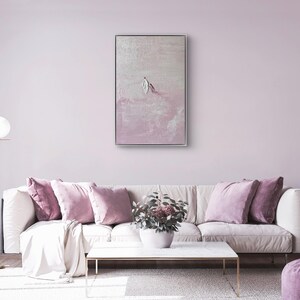 Pode incluir: Um interior de sala de estar com um sof&aacute; branco adornado com almofadas malva e um pufe a condizer. Uma pintura abstrata emoldurada est&aacute; pendurada acima do sof&aacute;. Uma lumin&aacute;ria de ch&atilde;o dourada e uma mesa de centro branca completam a decora&ccedil;&atilde;o. O quarto &eacute; pintado em um tom rosa suave.
