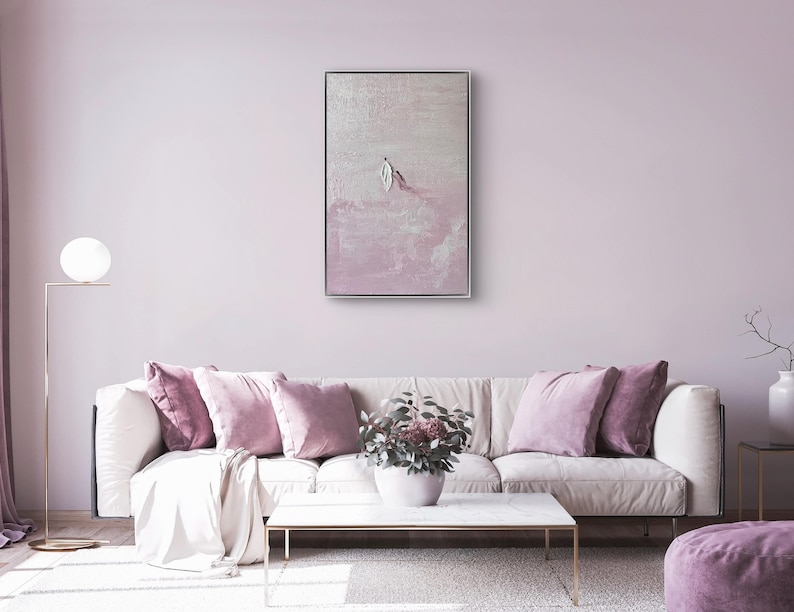 Pode incluir: Um interior de sala de estar com um sof&aacute; branco adornado com almofadas malva e um pufe a condizer. Uma pintura abstrata emoldurada est&aacute; pendurada acima do sof&aacute;. Uma lumin&aacute;ria de ch&atilde;o dourada e uma mesa de centro branca completam a decora&ccedil;&atilde;o. O quarto &eacute; pintado em um tom rosa suave.