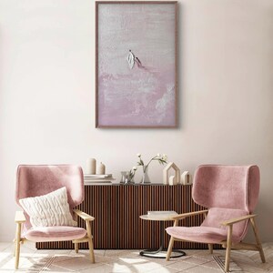 Pode incluir: Obra de arte abstrata emoldurada em madeira, com um design texturizado em tons de rosa e branco. A pintura retrata duas figuras, uma com forma de pena. A obra est&aacute; pendurada acima de duas poltronas rosa e um m&oacute;vel de madeira.