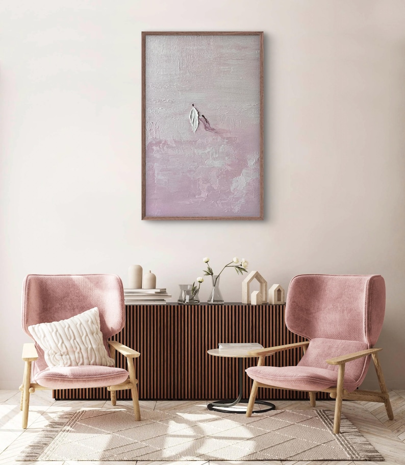 Pode incluir: Obra de arte abstrata emoldurada em madeira, com um design texturizado em tons de rosa e branco. A pintura retrata duas figuras, uma com forma de pena. A obra est&aacute; pendurada acima de duas poltronas rosa e um m&oacute;vel de madeira.