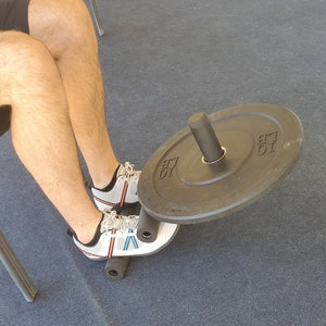 May include: A person's legs are bent at the knee with a weight plate on a bar resting on their lower legs. The weight plate is black with white lettering that says "10 LB".