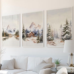 Könnte beinhalten: Drei gerahmte Aquarellbilder von verschneiten Berglandschaften mit Kiefern und einer kleinen Hütte. Die Bilder hängen an einer weißen Wand in einem Wohnzimmer mit einem weißen Sofa, einem weißen Sessel und einem schwarzen Beistelltisch.