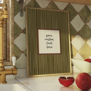 Puede incluir: Escena de cocina con un marco con el texto "your custom text here", un grifo dorado y un cuenco con granadas. El marco tiene un diseño a rayas verdes. Tazas y vasos blancos sobre una estantería de madera.