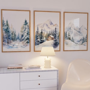 May include: Three framed watercolour paintings of winter landscapes with snowy mountains, trees, and a small cabin. The paintings are hung on a white wall above a white dresser with a lamp on top.