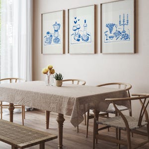 May include: Three framed blue and white illustrations of a table setting with food, wine, and candles. The illustrations are hanging on a light beige wall above a wooden table with a light beige tablecloth and four chairs.