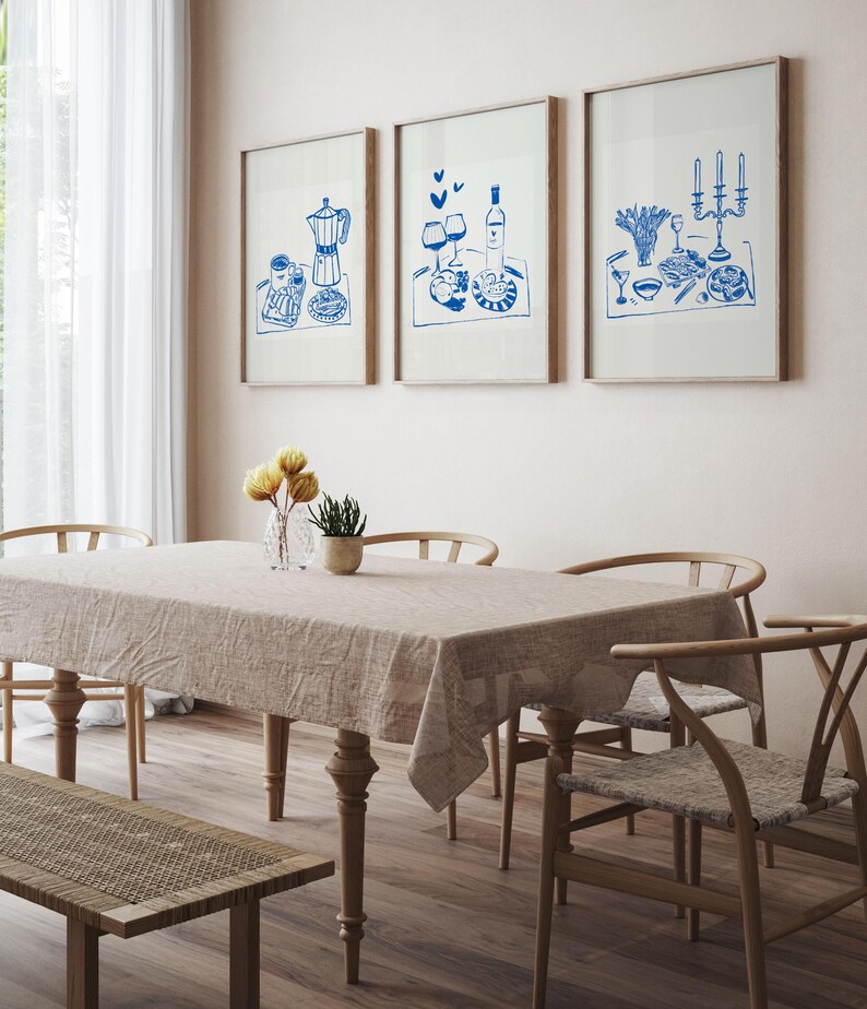 May include: Three framed blue and white illustrations of a table setting with food, wine, and candles. The illustrations are hanging on a light beige wall above a wooden table with a light beige tablecloth and four chairs.