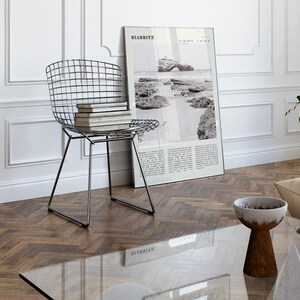 Pu&ograve; includere: Un moderno soggiorno con una sedia in filo metallico con libri, una fotografia in bianco e nero incorniciata e un tavolino con piano in vetro. La fotografia &egrave; di Biarritz, Francia. La stanza ha un pavimento in legno e pareti bianche.