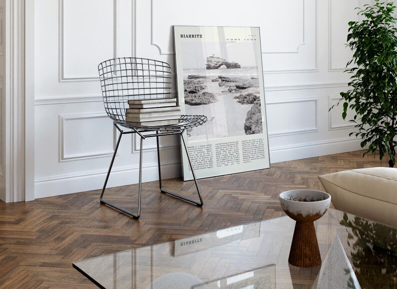 Pu&ograve; includere: Un moderno soggiorno con una sedia in filo metallico con libri, una fotografia in bianco e nero incorniciata e un tavolino con piano in vetro. La fotografia &egrave; di Biarritz, Francia. La stanza ha un pavimento in legno e pareti bianche.