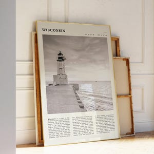 May include: A framed black and white print featuring a lighthouse on a pier, with the word "Wisconsin" at the top. The print has a gold frame and is leaning against a white wall. The print includes text describing Wisconsin.