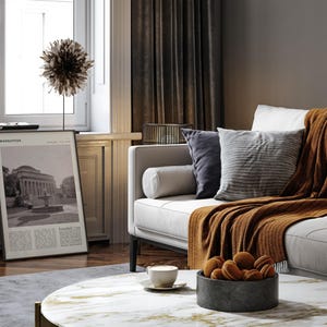 May include: A living room interior with a white sofa, cushions, and a rust-coloured throw. A framed print with the word "Manhattan" is visible. A round coffee table with a marble top holds a bowl of biscuits and a cup of coffee.