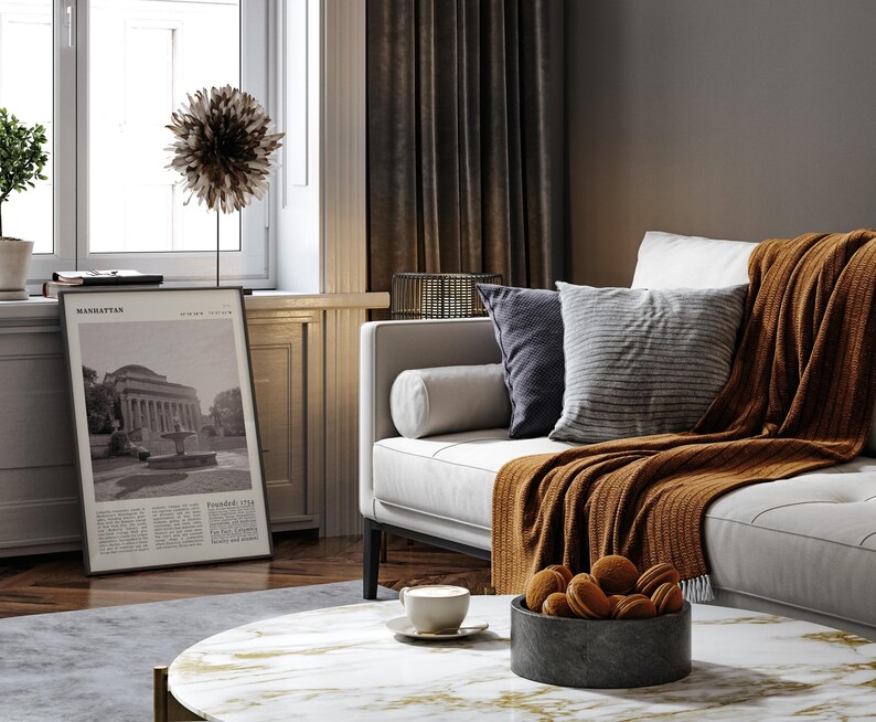 May include: A living room interior with a white sofa, cushions, and a rust-coloured throw. A framed print with the word "Manhattan" is visible. A round coffee table with a marble top holds a bowl of biscuits and a cup of coffee.