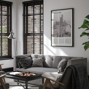 K&ouml;nnte beinhalten: Wohnzimmerinterieur mit grauem Sofa, Kissen und gemusterter Decke. Ein schwarz-wei&szlig; gerahmter Druck mit Text und Stadtansicht h&auml;ngt an der Wand. Dunkle Holzjalousien bedecken die Fenster.