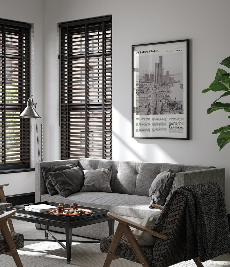 K&ouml;nnte beinhalten: Wohnzimmerinterieur mit grauem Sofa, Kissen und gemusterter Decke. Ein schwarz-wei&szlig; gerahmter Druck mit Text und Stadtansicht h&auml;ngt an der Wand. Dunkle Holzjalousien bedecken die Fenster.