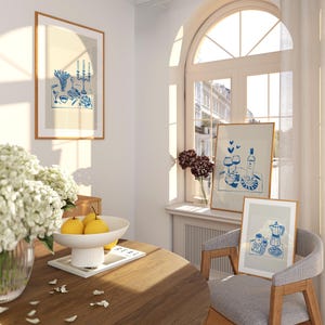 May include: A dining room with a wooden table, two chairs, and three framed illustrations. The illustrations are in a blue and white color scheme and depict a table setting, a wine bottle and glasses, and a coffee pot and cups. The table is set with a white bowl of fruit and a magazine.