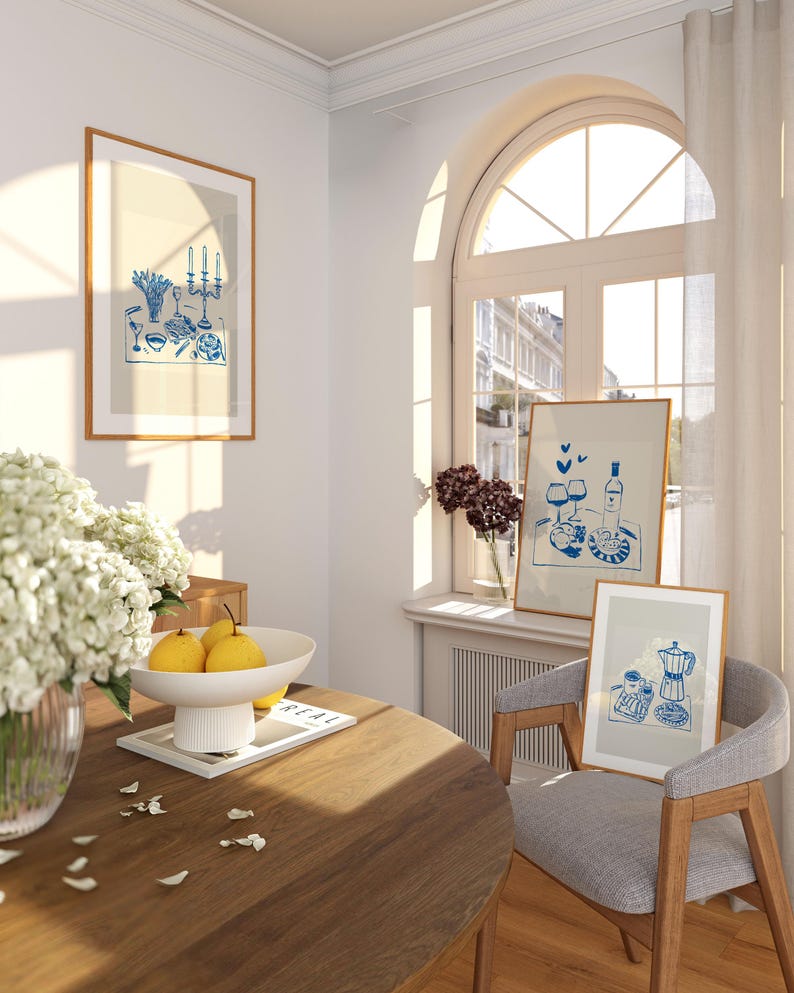 May include: A dining room with a wooden table, two chairs, and three framed illustrations. The illustrations are in a blue and white color scheme and depict a table setting, a wine bottle and glasses, and a coffee pot and cups. The table is set with a white bowl of fruit and a magazine.