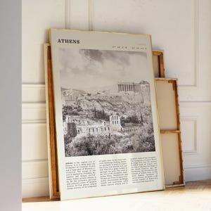 May include: A framed black and white print of Athens, Greece, featuring the Acropolis and surrounding buildings. The print has a gold frame and is leaning against a white wall. The word "Athens" is at the top.