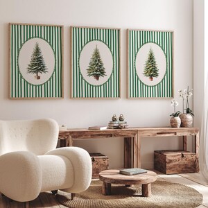 May include: Three framed prints featuring Christmas trees in oval frames against a green and white striped background. The frames are a natural wood color. The prints are displayed above a wooden console table with a white chair in the foreground.