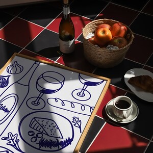 May include: A framed white drawing of a cheese plate with wine glasses, a baguette, and grapes. The drawing is on a table with a bottle of wine, a bowl of apples, and a cup of coffee.