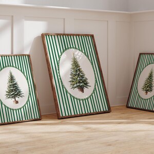 May include: Three framed art prints featuring a Christmas tree illustration. Each print has a brown wooden frame, a green and white striped background, and an oval-shaped tree image. The prints are leaning against a white wall and a wooden floor.