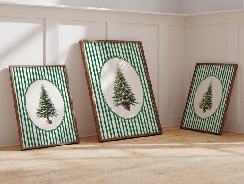 May include: Three framed art prints featuring a Christmas tree illustration. Each print has a brown wooden frame, a green and white striped background, and an oval-shaped tree image. The prints are leaning against a white wall and a wooden floor.