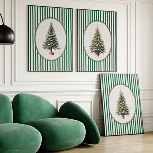 May include: Three framed Christmas tree art prints. The prints feature a green and white striped background with an oval-shaped image of a Christmas tree. The frames are black. A green velvet sofa is in the foreground.