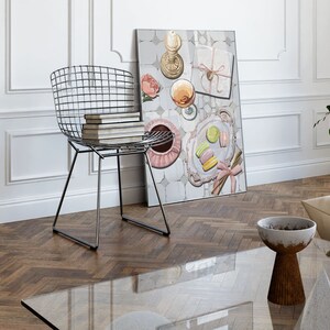 May include: A framed print of a still life featuring a pink and white floral design, a glass of wine, a plate of macarons, and a gift box with a pink ribbon. The print is hanging on a white wall above a glass coffee table and a metal wire chair.