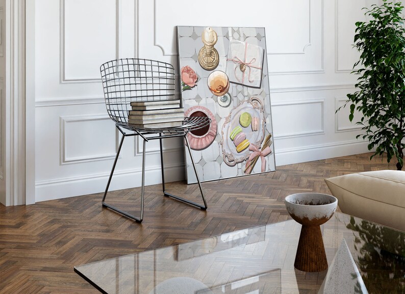 May include: A framed print of a still life featuring a pink and white floral design, a glass of wine, a plate of macarons, and a gift box with a pink ribbon. The print is hanging on a white wall above a glass coffee table and a metal wire chair.