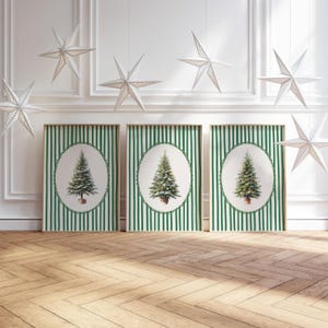 May include: Three framed Christmas tree prints with green and white striped backgrounds. Each print features a watercolor-style Christmas tree in an oval frame. White star-shaped decorations hang from the ceiling. The floor is made of wood.